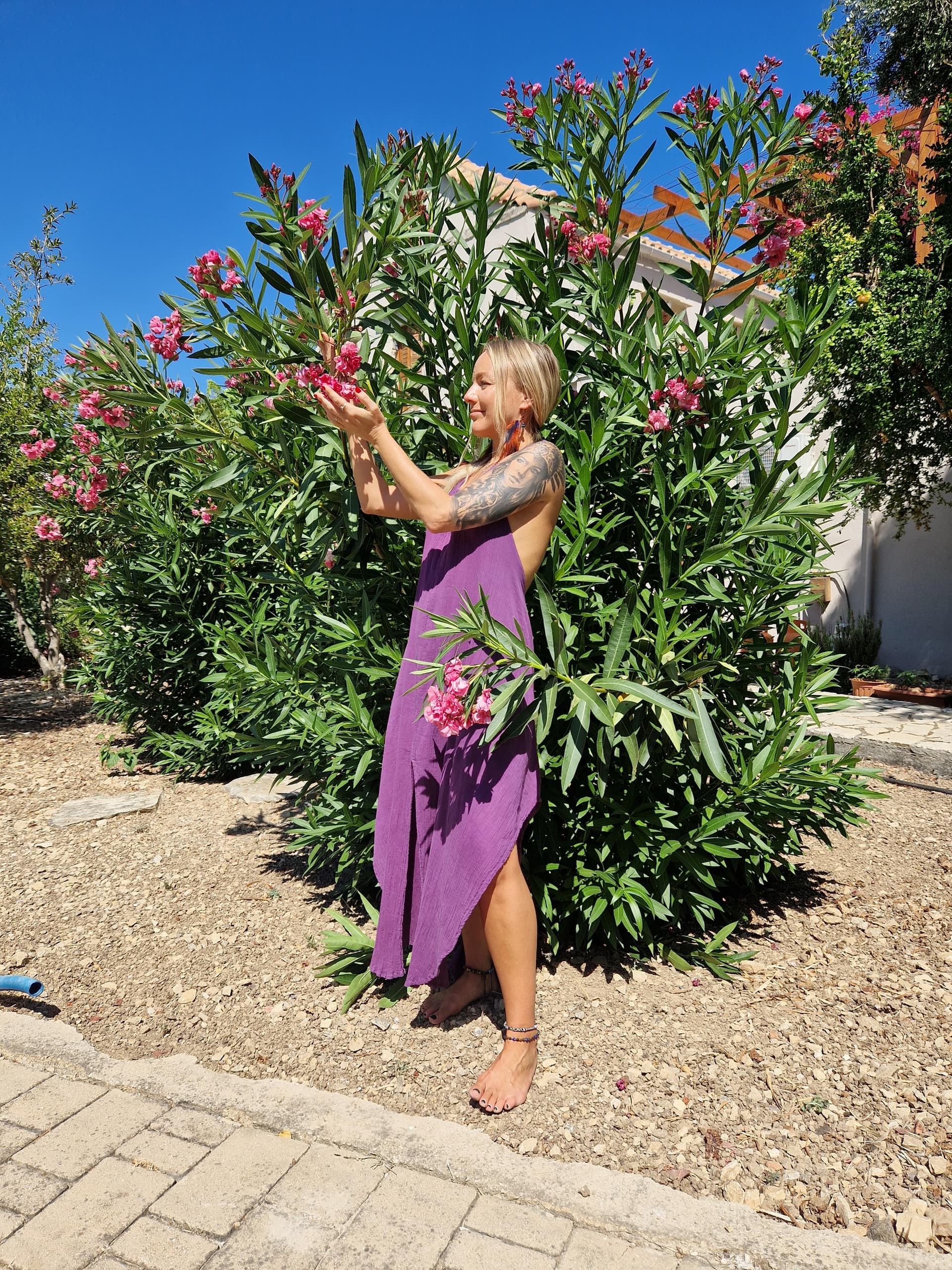 Lina in a purple dress reaching for pink flowers in a Mediterranean garden